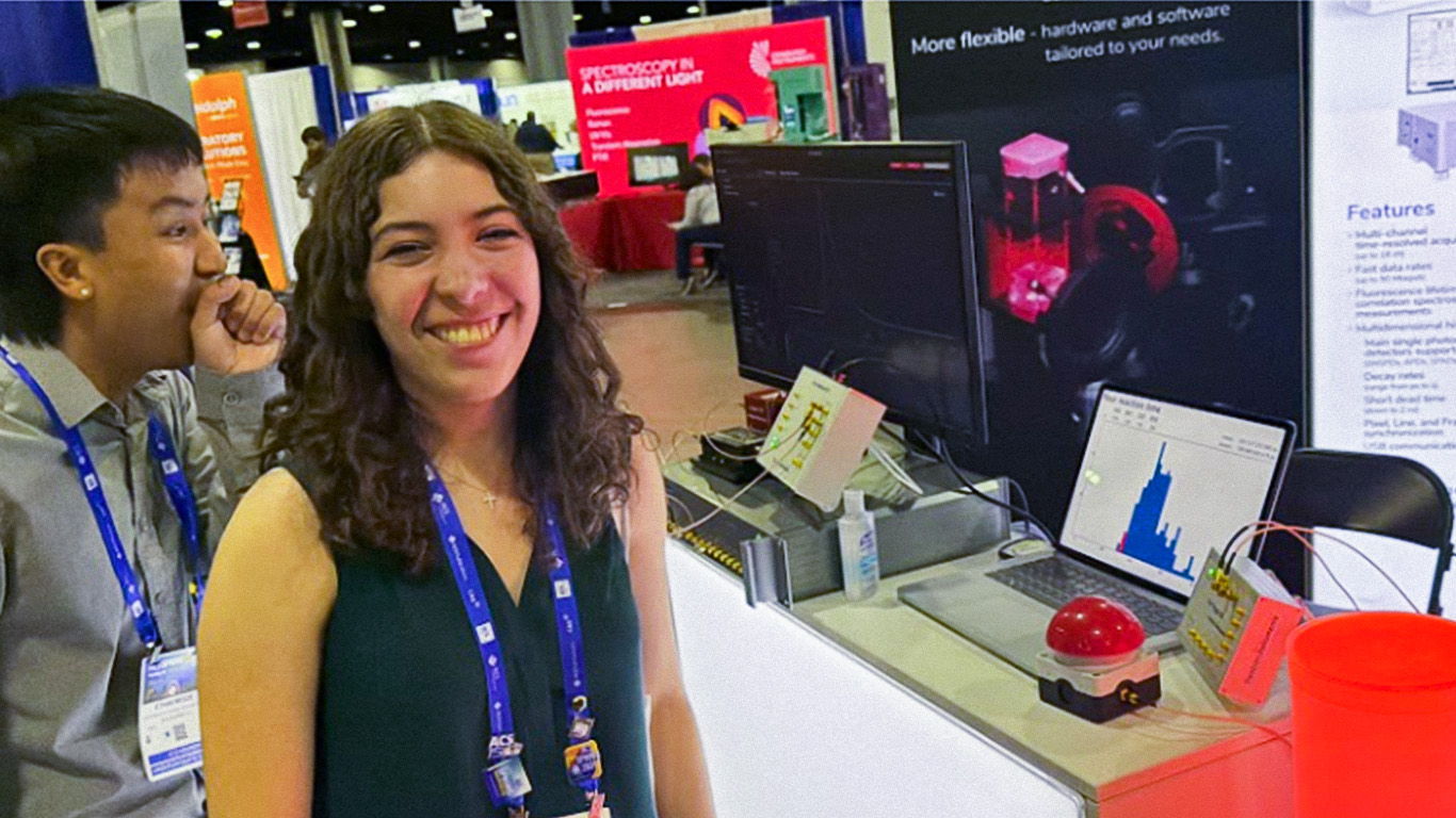 Two students at Booth 805 at ACS Spring 2026 in Atlanta, one smiling while playing the Swabian Instruments reaction-time game. The booth backdrop highlights Dynamic Light Scattering and TCSPC. A laptop displaying a live reaction-time histogram is visible on the counter, alongside the red-button game setup and DLScat instrumentation.