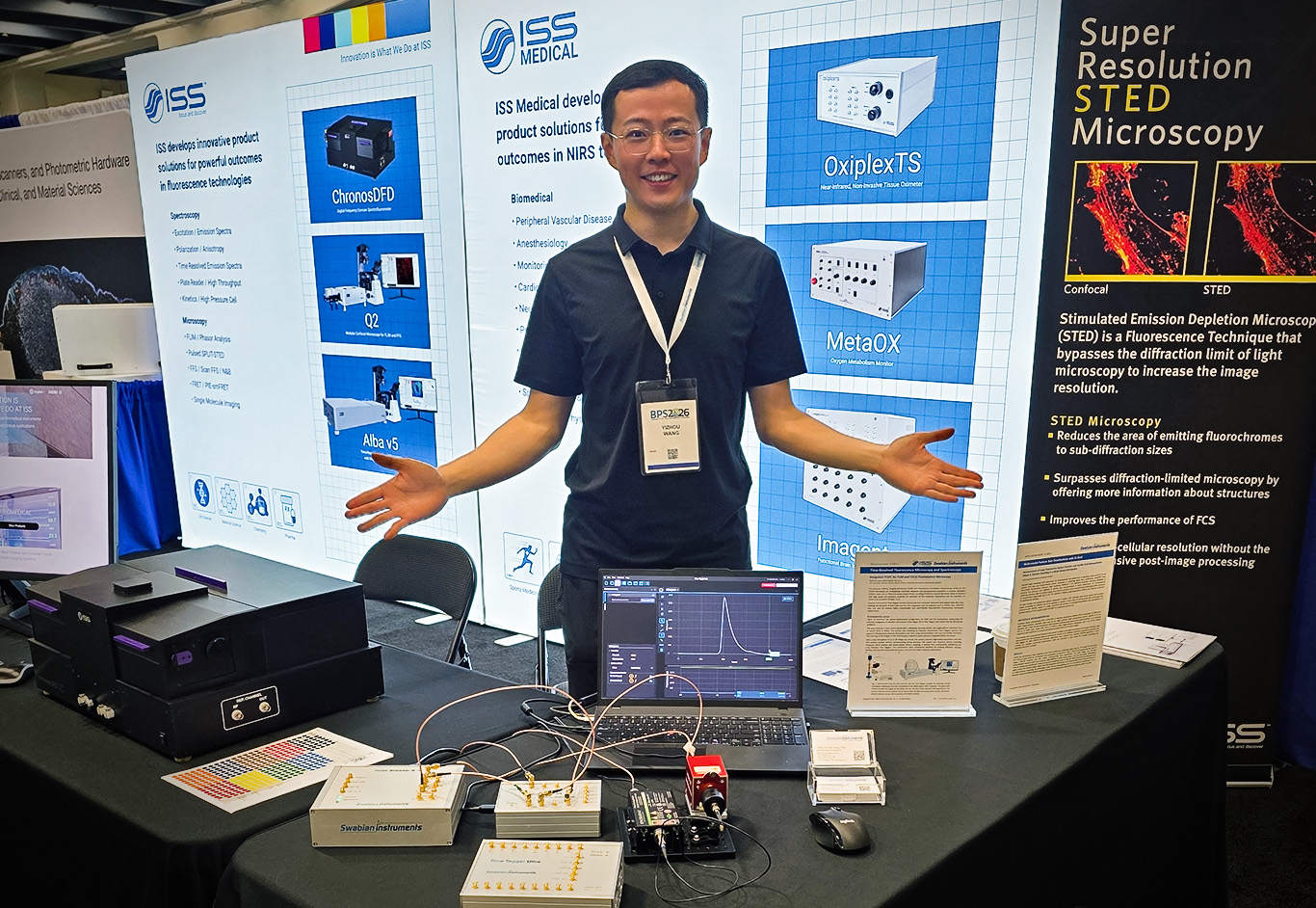 Photo of Swabian Instruments team member Yizhou Wang at the joint Swabian Instruments and ISS booth during the BPS Meeting 2026 in San Francisco. He is demonstrating a fluorescence lifetime measurement setup featuring a laser, Pulse Streamer, Time Tagger, and SPAD detector, while the laptop screen displays the fluorescence lifetime signal.