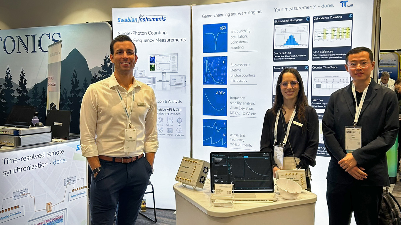 Swabian Instruments team members at one of the exhibition booths for BiOS, standing behind a monitor that displays a fluorescence lifetime measurement using a Time Tagger. The team is standing in front of the informational walls that explain the measurements possible with the Time Tagger Series. From left to right: Thomas Hayes, Mireia Perera Gonzalez, Yizhou Wang.