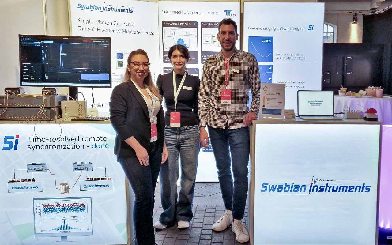 Swabian Instruments team standing in front of their exhibition booth at European Quantum Technologies Conference 2025. The booth displays the TimeTagger X, Pulse Streamer, and TimeTagger Ultra as part of a full White Rabbit pico-second synchronisation setup. The team is positioned beside the interactive Red Button game.