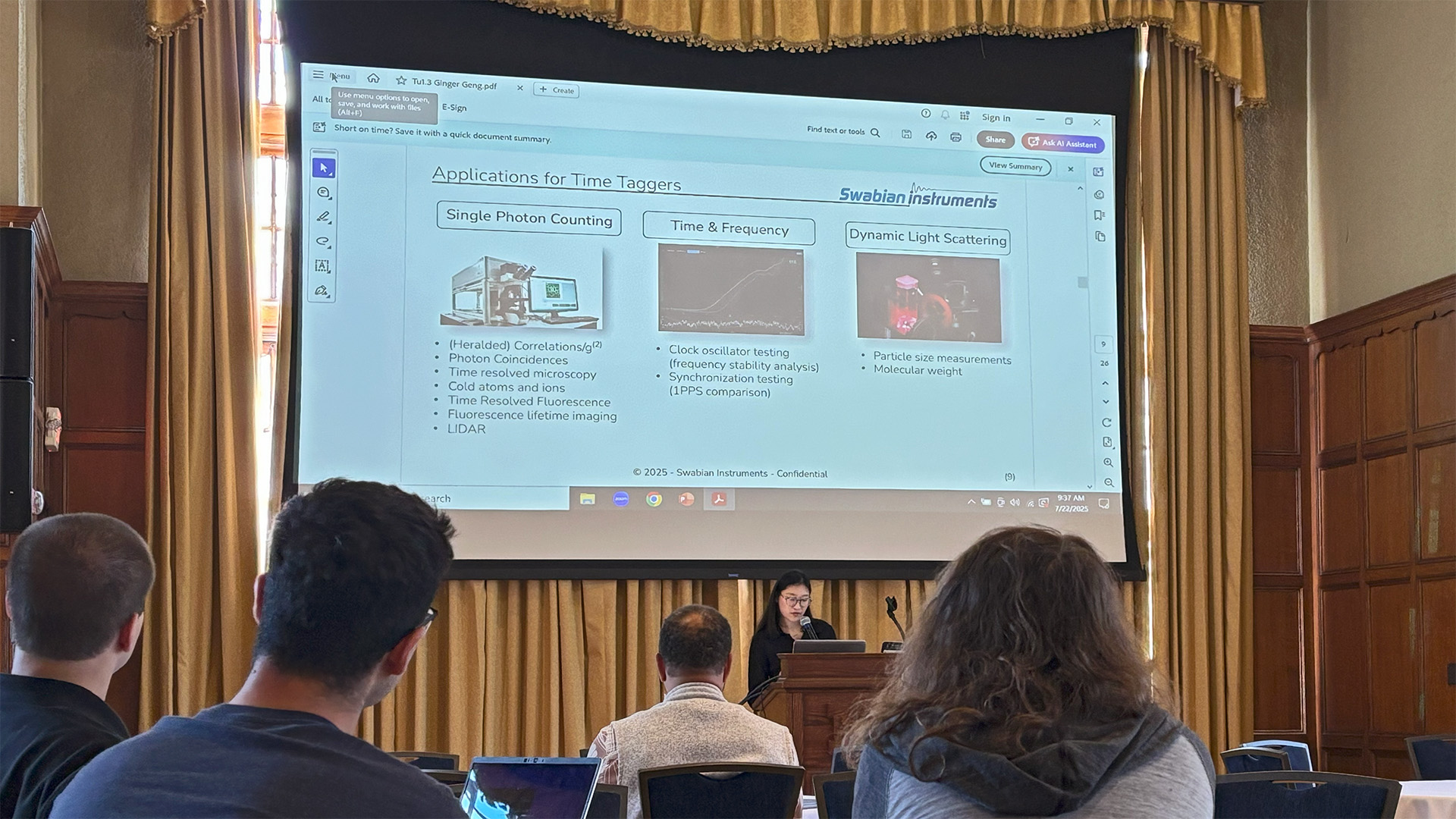 Ginger Geng from Swabian Instruments stands at a podium, presenting on High-Precision Time Tagging for Quantum Technologies at MQC Entanglement 2025. The presentation slide summarizes applications for Time Taggers, listing uses in Single Photon Counting, Time & Frequency, and Dynamic Light Scattering.