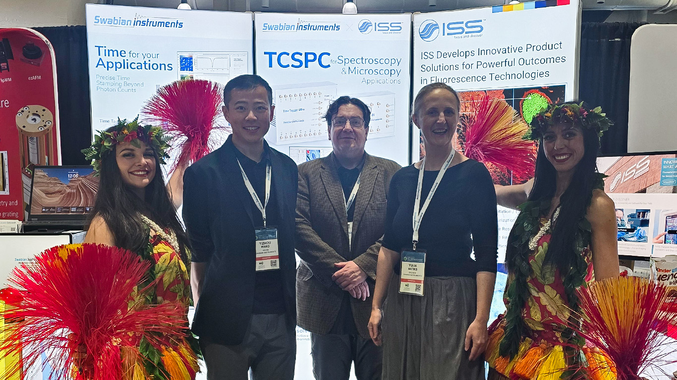 Group photo at the Swabian Instruments and ISS booth (#804) during MRS Fall Meeting 2025 in Boston, showing Yizhou Wang, James Keeler (ISS), and Yulia Matiks standing in front of the booth. Two MRS staff members wearing Hawaiian skirts stand nearby, indicating that the next MRS meeting will be held in Hawaii.