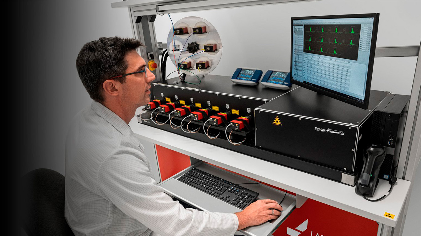A person using the laboratory workstation showing a detector characterization setup on a ThorLabs optical bench. The rack contains six COUNT detector modules with fiber connections, flanked by a Swabian Instruments Time Tagger Ultra unit. Two small display monitors sit on top of the rack, and a computer monitor displays timing histograms. A circular mount holding several detectors is visible on the left, and four laser driver modules are mounted on the upper rail.
